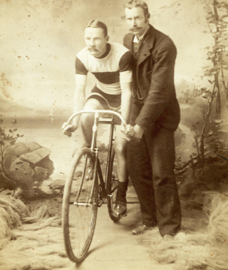 An 1896 studio photo shows Percy Armstrong in period cycling attire with a moustache that matches his handlebars, mounted on a racing bicycle held by his trainer Mr. DM Leslie.