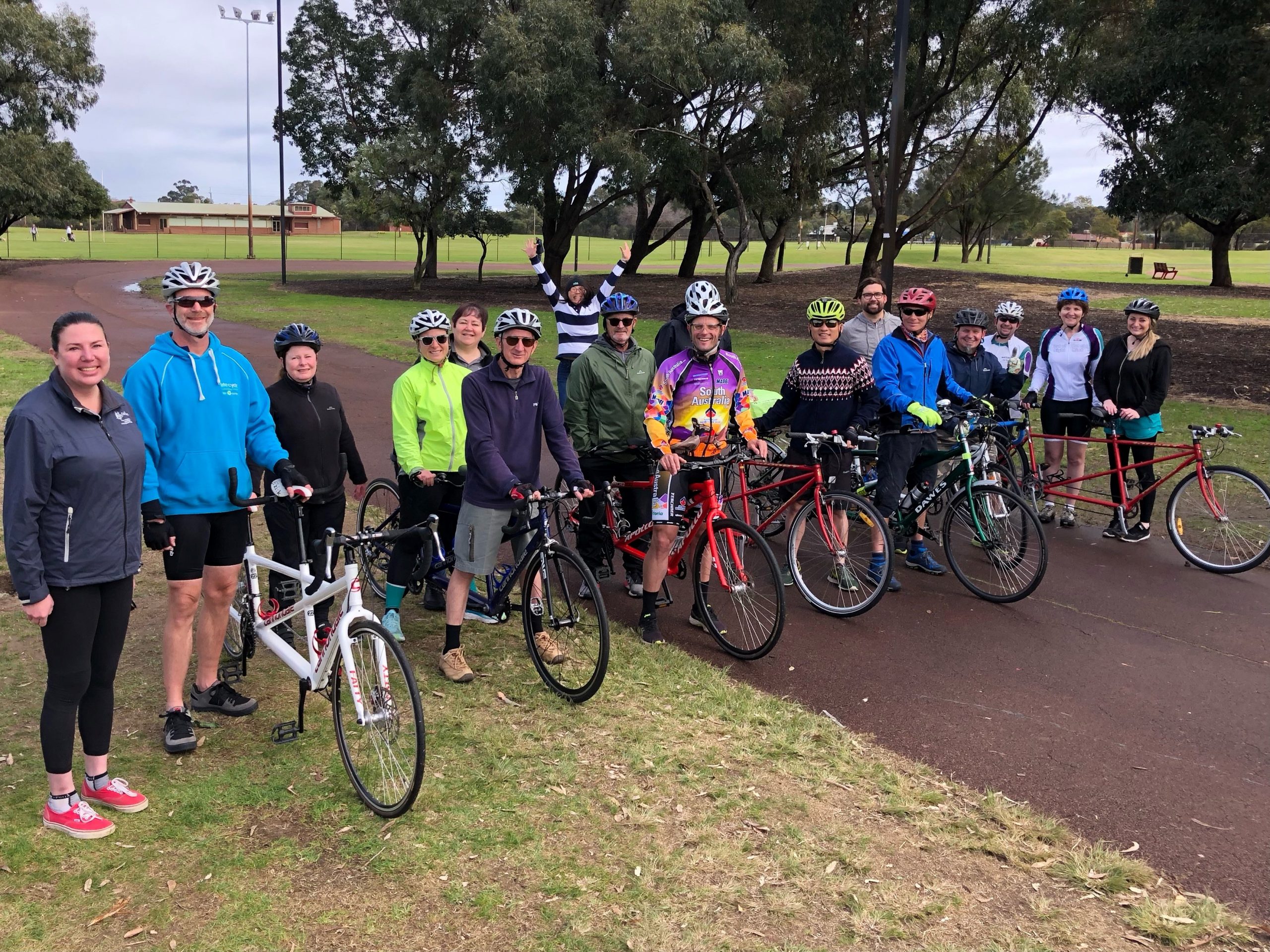 A group of 17 people stand together with 7 tandem bikes for a group photo while one member celebrates with a start jump at the back of the group. Oh what a feeling!