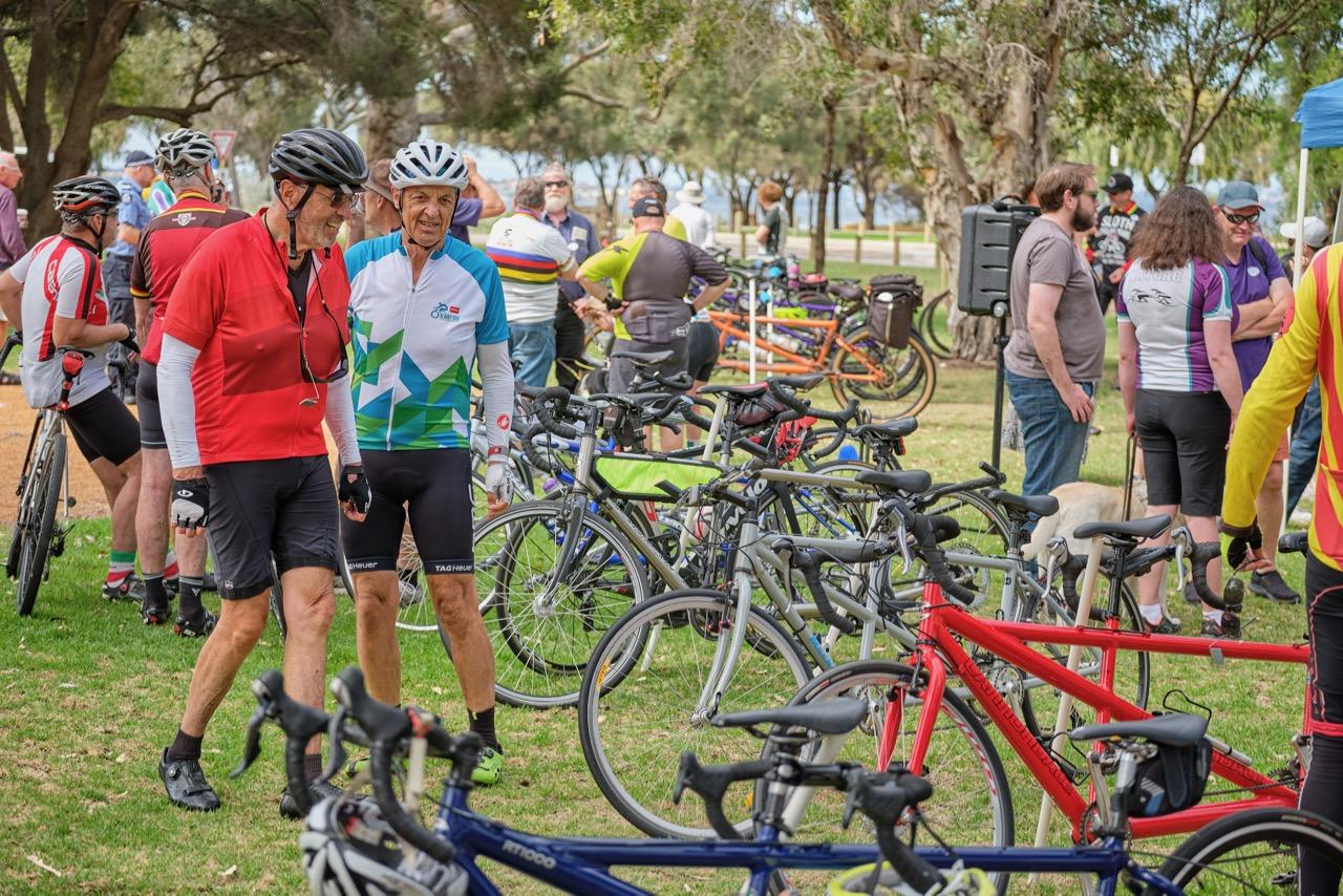 Image shows a grassed area with a long row of tandem bicycles and people nearby chatting.