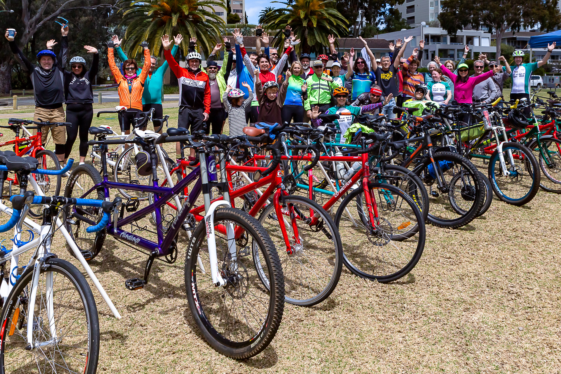 Around 30-40 tandem cyclists of all ages are standing and waving with arms raised, around 20 tandem bikes of all shapes, sizes and colours are lined up in the foreground.