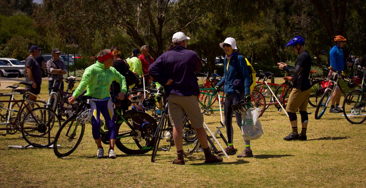 People are standing around the collection of tandem bicycles spread across the foreshore lawns.