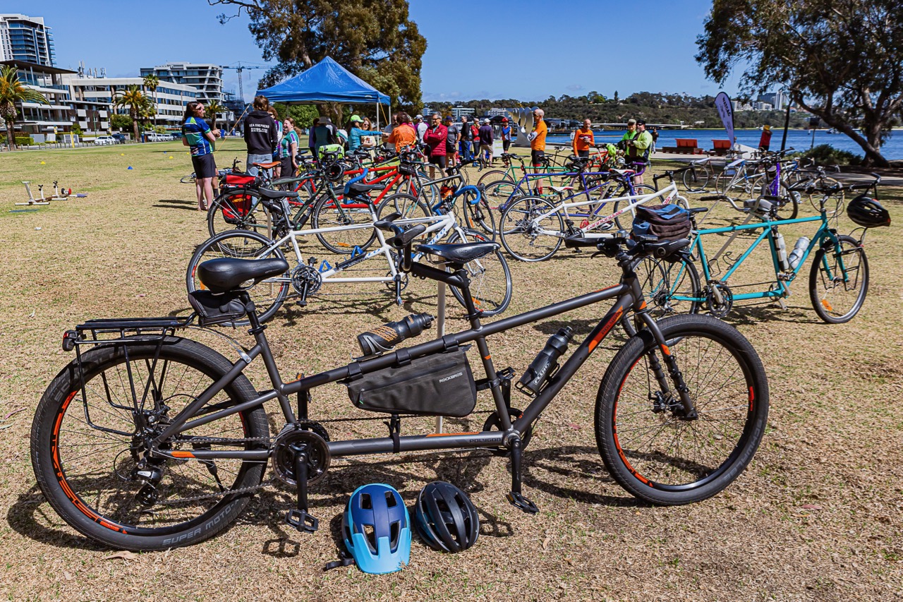 People stand chatting behind a large array of tandem bikes lined up on the lawn of the South Perth foreshore.