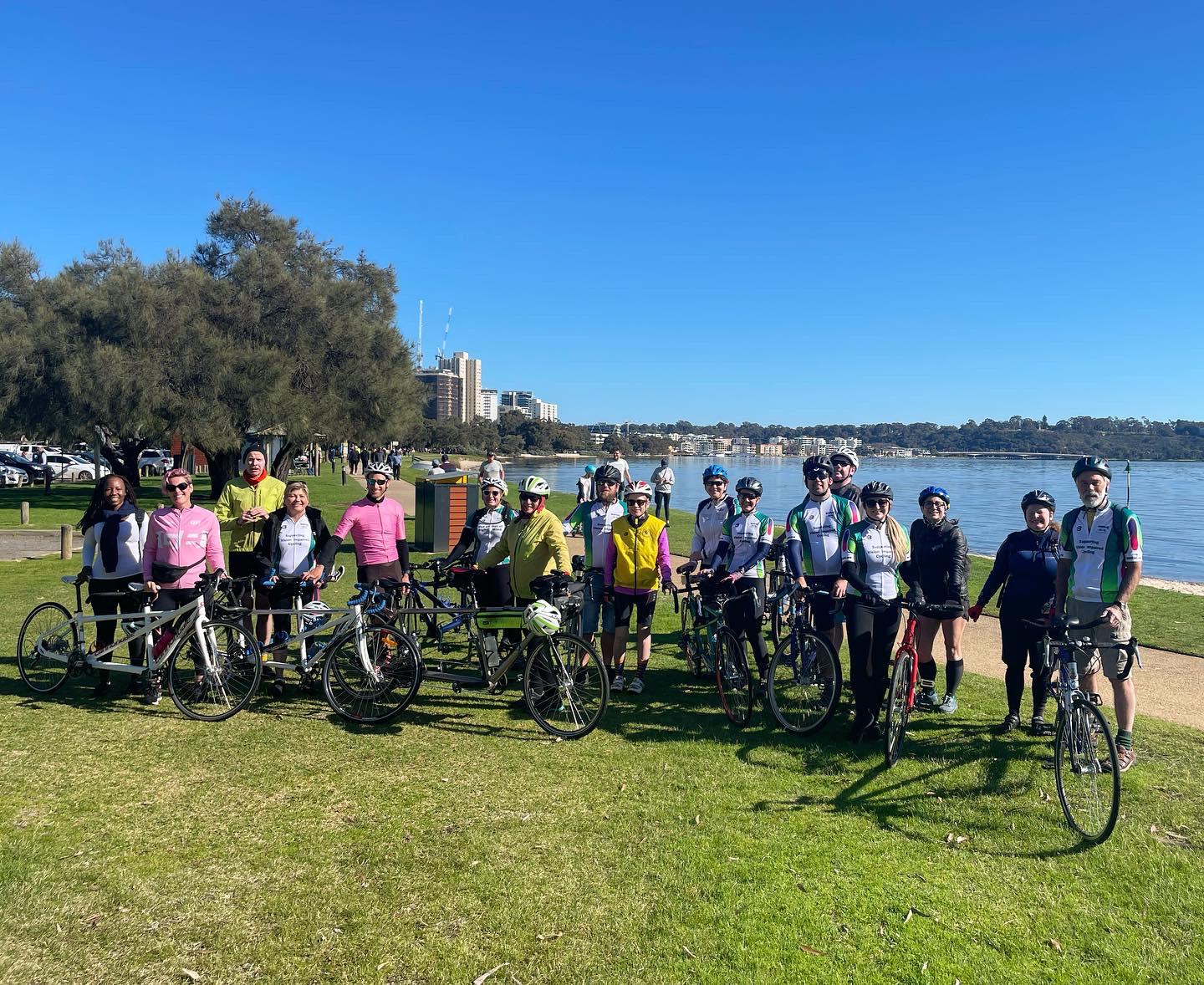 Nineteen people are standing on the South Perth foreshore with 9 tandems and a single bike lined up on the grass.  The group included 8 blind or vision-impaired stokers, two of whom enjoyed their first ride with WATCAC.