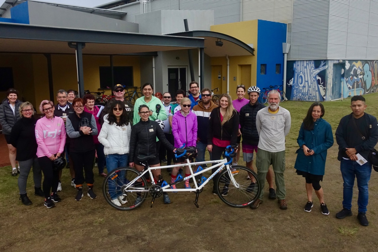 Twenty two people are grouped together around a tandem cycle, standing adjacent to the Leisure Centre in Karawara.